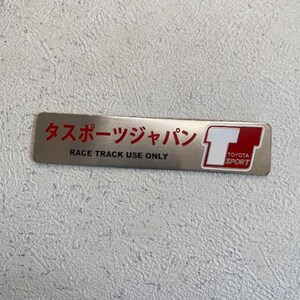May include: A rectangular, brushed metal emblem with red Japanese text and the English text "RACE TRACK USE ONLY." The right side features a white "T" inside a red square, with "TOYOTA SPORT" below.