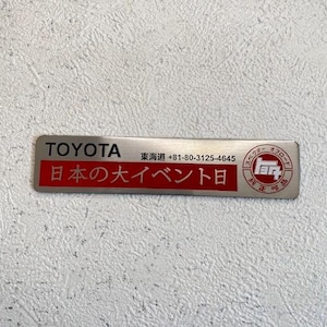 Op de afbeelding: Een rechthoekige zilverkleurige metalen badge met het woord "TOYOTA" bovenaan. Daaronder staat een rode rechthoek met Japanse tekst. Aanvullende Japanse tekst en een logo staan aan de rechterkant. De badge bevat ook een telefoonnummer.