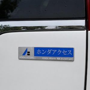May include: A rectangular silver and blue metal emblem attached to a white vehicle surface. The emblem features the Honda Access logo, the text "HONDA ACCESS", Japanese characters, and the address "HONDA Niiza-shi 81 4 8477 8217" in white.