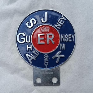 May include: A vintage blue and red metal badge with the text "Guernsey" and "Alderney" in white lettering. The badge has a central red circle with "ER" and a crown. A silver metal tab with two holes is attached.