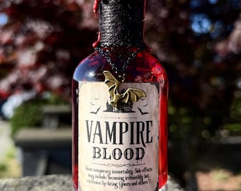 Puede incluir: Una botella de vidrio llena de líquido rojo, etiquetada como "VAMPIRE BLOOD". El cuello de la botella está envuelto con cuerda negra y sellado con cera roja. Un colgante de murciélago cuelga del cuello. Un artículo único de Halloween.