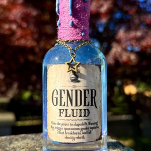 Può includere: Una bottiglia di vetro decorativa riempita di liquido blu, etichettata "GENDER FLUID". La bottiglia ha un collo avvolto in rosa, gocce di cera blu e un ciondolo a forma di stella dorata. L'etichetta include testo sulla metamorfosi e la rinascita dell'identità.