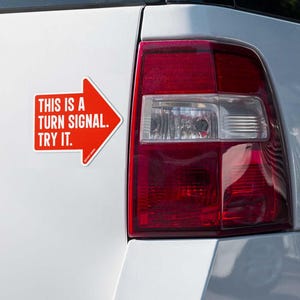 Puede incluir: Una pegatina roja en forma de flecha en la parte trasera de un vehículo blanco. La pegatina tiene el texto blanco: "THIS IS A TURN SIGNAL. TRY IT." El piloto trasero del vehículo es visible.