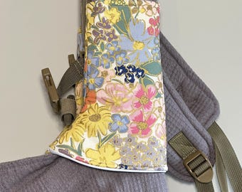 May include: A floral print baby carrier cover with a white trim. The cover features a colorful pattern of flowers in yellow, pink, blue, and gold. The carrier is attached to a gray waffle-knit fabric with olive green straps and buckles.