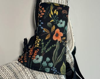 May include: A black floral print baby carrier cover with orange, yellow, and blue flowers. The cover is attached to a black and white patterned baby carrier. The cover is designed to protect the baby carrier from the elements.