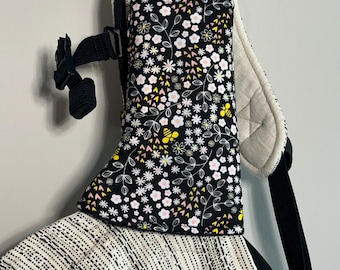 May include: A black and white patterned baby carrier with a floral print pocket. The pocket features white, pink, and yellow flowers, along with yellow bees. The carrier has black straps and a black buckle.