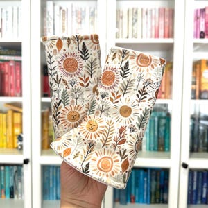 May include: Two fabric book sleeves with a repeating pattern of smiling suns, leaves, and floral elements in shades of orange, brown, and green on a cream background. The sleeves are held up against a white bookshelf.