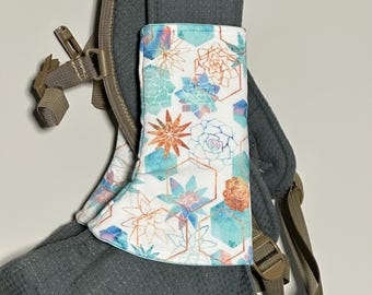 May include: A white and patterned baby carrier cover with a succulent design in teal, orange, and blue. The cover is attached to a gray baby carrier with brown straps and buckles.