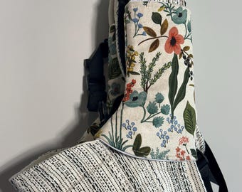 May include: A baby carrier with a floral pattern in shades of red, blue, and green on a cream background. The carrier has a black and white woven fabric and black straps and buckles.