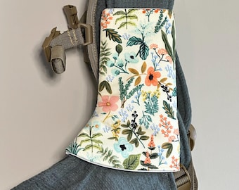 May include: A cream-colored fabric strap cover featuring a colorful botanical print of various flowers, leaves, and berries in shades of orange, pink, blue, and green, attached to a dark gray textured baby carrier strap with brown plastic buckles.