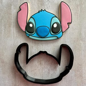 Pode incluir: Um biscoito decorado em forma de cabeça de um personagem alienígena azul com grandes orelhas cor-de-rosa, olhos pretos e nariz azul, posicionado acima de um cortador de biscoitos de plástico preto que corresponde ao contorno da cabeça e orelhas.