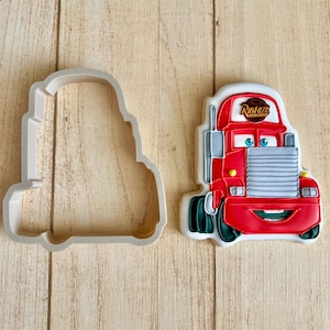 Peut inclure: Un emporte-pièce en forme de camion rouge et un biscuit décoré assorti représentant un camion rouge avec une calandre grise, des yeux bleus et un panneau blanc sur le dessus avec le texte Rust-eze. Le biscuit a des roues vert foncé.