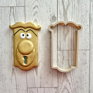 May include: A gold-colored cookie shaped like a door handle with a face, featuring two white eyes with black pupils and a keyhole, positioned next to a matching beige plastic cookie cutter outline of the same door handle shape.