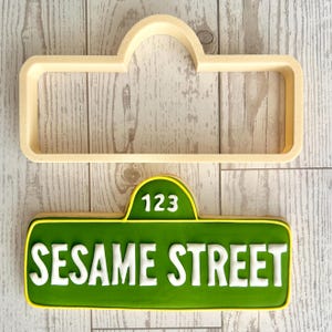 May include: A beige plastic cookie cutter shaped like a street sign and a corresponding green cookie decorated with white icing that reads 123 SESAME STREET. The cookie has a rectangular base with a rounded top section and a yellow border.