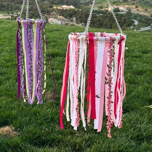 May include: Two hanging decorative wind chimes. One features purple and green ribbons, the other pink, white, and floral ribbons. Both are suspended by silver chains and set against a green grass background.