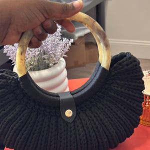 May include: Black crochet handbag with a circular handle made of light brown material. The bag has a black leather strap with a gold-colored button closure. The bag is sitting on a red surface.