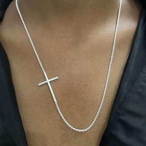 Może przedstawiać: Srebrny naszyjnik z boczną zawieszką w kształcie krzyża. Krzyż jest przymocowany do delikatnego łańcuszka. Naszyjnik jest prezentowany na ciemnym tle.