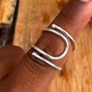 May include: A silver-colored ring with a unique, open design. The ring wraps around the finger with a curved shape and small, rounded ends. The ring is made of a shiny, metallic material.