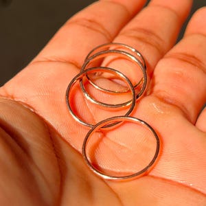 Op de afbeelding: Een hand die verschillende dunne, goudkleurige ringen vasthoudt. De ringen zijn eenvoudige banden, waarschijnlijk van metaal, en zijn op elkaar gestapeld. De belichting benadrukt de metalen glans van de ringen, wat een warme uitstraling creëert. De ringen zijn een minimalistisch sieraad.