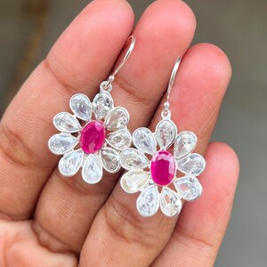 May include: A pair of silver flower earrings. Each earring has a vibrant, oval ruby centre, encircled by clear, petal-shaped stones. The earrings are held by a person's hand, highlighting their delicate design and craftsmanship.