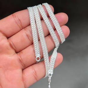 Peut inclure: Un collier en argent avec un motif tissé en forme de maille. Le collier est tenu dans une main, mettant en valeur sa texture détaillée et la façon dont il capte la lumière. Il possède une fermeture à fermoir.
