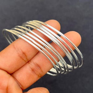 Könnte beinhalten: Eine Sammlung dünner, silberfarbener Armreifen. Die Armreifen sind übereinander gestapelt, wodurch ein geschichteter Effekt entsteht. Die glatte, metallische Oberfläche reflektiert das Licht und verleiht ihnen ein helles Aussehen. Die Armreifen werden vor einem dunklen Hintergrund gehalten.