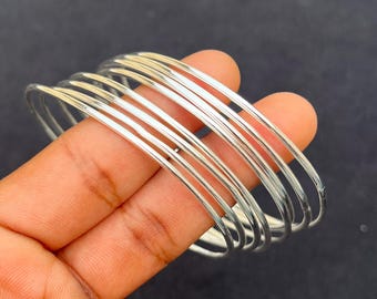 May include: A collection of thin, silver-colored bangles. The bangles are stacked together, creating a layered effect. The smooth, metallic surface reflects light, giving them a bright appearance. The bangles are held against a dark background.