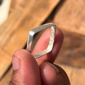 Puede incluir: Un anillo plateado de forma cuadrada con esquinas redondeadas. El anillo se sostiene contra un fondo de madera borroso. El diseño del anillo es simple y moderno, adecuado para el uso diario. El anillo es una joya.