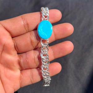 Op de afbeelding: Een zilveren armband met een heldere turquoise ovale steen gezet in een zilveren schakelketting ontwerp. De turquoise steen heeft een levendige blauwe kleur en de armband heeft een gepolijste afwerking.