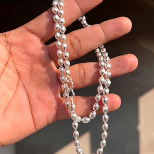 Puede incluir: Un collar plateado con una serie de cuentas ovaladas. El collar está hecho de un material metálico brillante y tiene un cierre de broche. Las cuentas están espaciadas uniformemente y crean un diseño simétrico. El collar se sostiene en una mano.