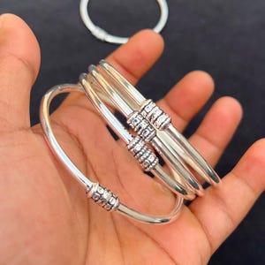 May include: A collection of silver bangles featuring decorative beaded detailing. The bangles have a smooth, polished finish and a simple, elegant design. The image shows several bangles, some stacked together, and one in the background.