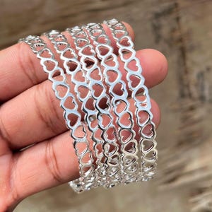 May include: A stack of silver bangles with heart-shaped cutouts. The bangles are made of a shiny metal and are stacked together. The heart cutouts create a delicate and romantic design.