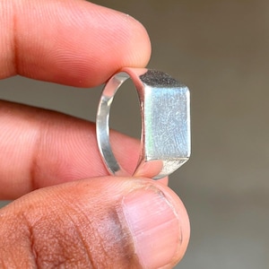 May include: A silver signet ring with a rectangular face. The ring is held between fingers, showcasing its polished surface. The ring's design is simple and modern, suitable for everyday wear.