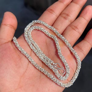 May include: A silver-colored chain necklace with a detailed, woven design. The necklace is displayed on a hand, showcasing its intricate links and a secure clasp. The chain appears to be made of metal.