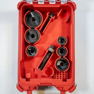May include: A red tool case with a clear lid, open to reveal various black tools. The tools include circular pieces and cylindrical components, neatly arranged within the case. The case is set against a white background.