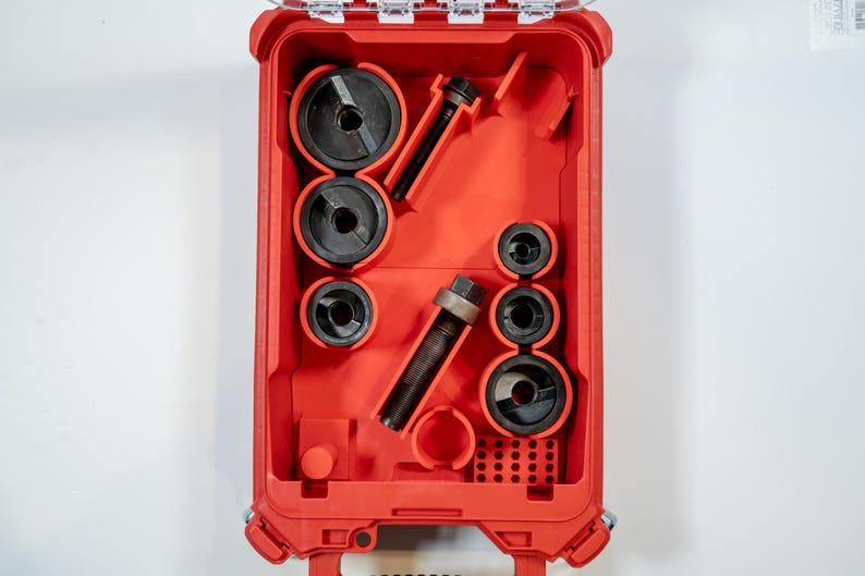 May include: A red tool case with a clear lid, open to reveal various black tools. The tools include circular pieces and cylindrical components, neatly arranged within the case. The case is set against a white background.
