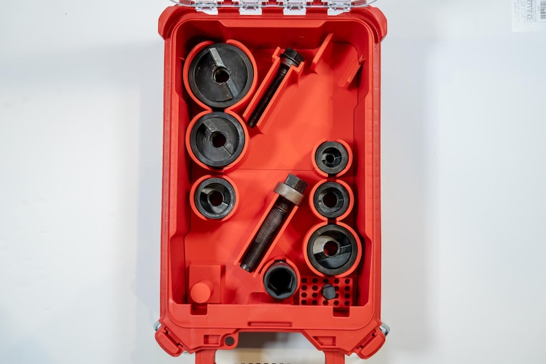 May include: A red tool case with a clear lid, filled with various black and silver metal tools. The tools include circular punch dies and bolts, neatly organized within the case. The case is designed for tool storage and organization.