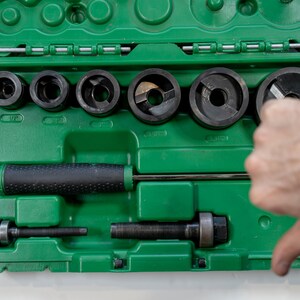 May include: A green toolbox with various black metal tools, including circular cutters and a handle. The tools are arranged inside the box, with a hand giving a thumbs-down gesture.