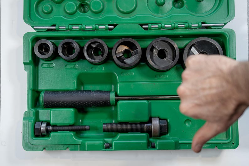 May include: A green toolbox with various black metal tools, including circular cutters and a handle. The tools are arranged inside the box, with a hand giving a thumbs-down gesture.