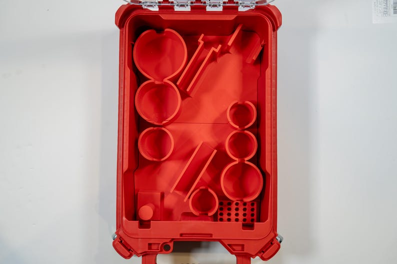 May include: Open red plastic toolbox with various compartments and molded shapes. The interior features circular and rectangular spaces, along with angled slots. The toolbox is designed for organizing tools and accessories.