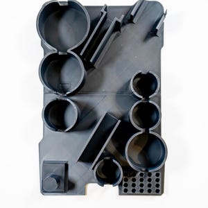 May include: A dark gray, rectangular organizer with various circular and rectangular compartments. The organizer has a series of cylindrical holes and angled slots. The design is modern and geometric, with a minimalist aesthetic.