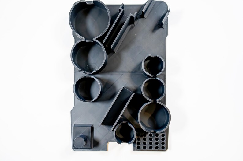 May include: A dark gray, rectangular organizer with various circular and rectangular compartments. The organizer has a series of cylindrical holes and angled slots. The design is modern and geometric, with a minimalist aesthetic.