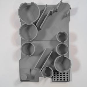 May include: A gray, molded plastic organizer with various compartments and shapes. It features circular holders, angled slots, and a small, rectangular section with multiple holes. The design is minimalist and functional.