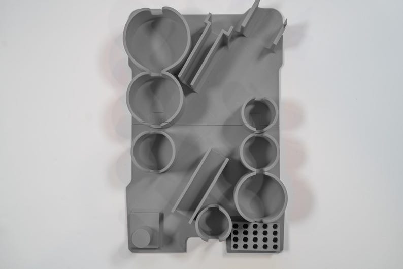 May include: A gray, molded plastic organizer with various compartments and shapes. It features circular holders, angled slots, and a small, rectangular section with multiple holes. The design is minimalist and functional.