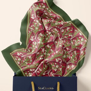 May include: A square silk scarf featuring a deep red background with a repeating pattern of white snowdrop flowers and green leaves, framed by a solid dark green border. The scarf is tucked into a navy blue shopping bag with the text SEACLAIRE.