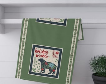 May include: A green kitchen towel featuring a repeating border of small pine trees and a central square graphic. The graphic displays a black bear silhouette decorated with floral patterns, red berries, and the red text holiday wishes.