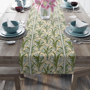 May include: A floral table runner with a repeating pattern of green leaves and blue flowers on a cream background. The runner is centered on a wooden table set for a meal, with plates, bowls, and glasses visible.