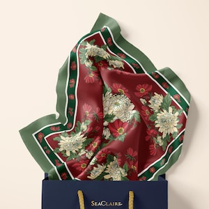 May include: A square silk scarf with a deep burgundy background featuring white chrysanthemum flowers and green leaves, framed by a dark green border. The scarf is tucked into a navy blue shopping bag with the gold text SeaClaire printed on it.