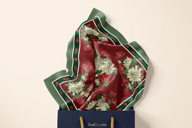 May include: A square silk scarf with a deep burgundy background featuring white chrysanthemum flowers and green leaves, framed by a dark green border. The scarf is tucked into a navy blue shopping bag with the gold text SeaClaire printed on it.