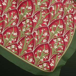 May include: A square fabric scarf featuring a repeating pattern of white snowdrop flowers with green leaves set against a deep red background. The design is organised into arched, scale-like segments outlined in white, finished with a solid green border.